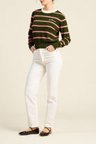 A model wearing a green crewneck sweater with pink horizontal stripes and a small white embroidered logo on the chest, paired with white pants and dark brown flat shoes, against a solid beige background.