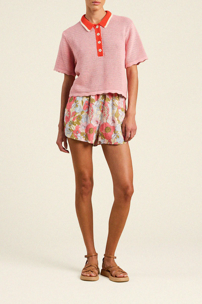 Person wearing a pink textured top with red collar and buttons, floral shorts, and sandals on a beige background