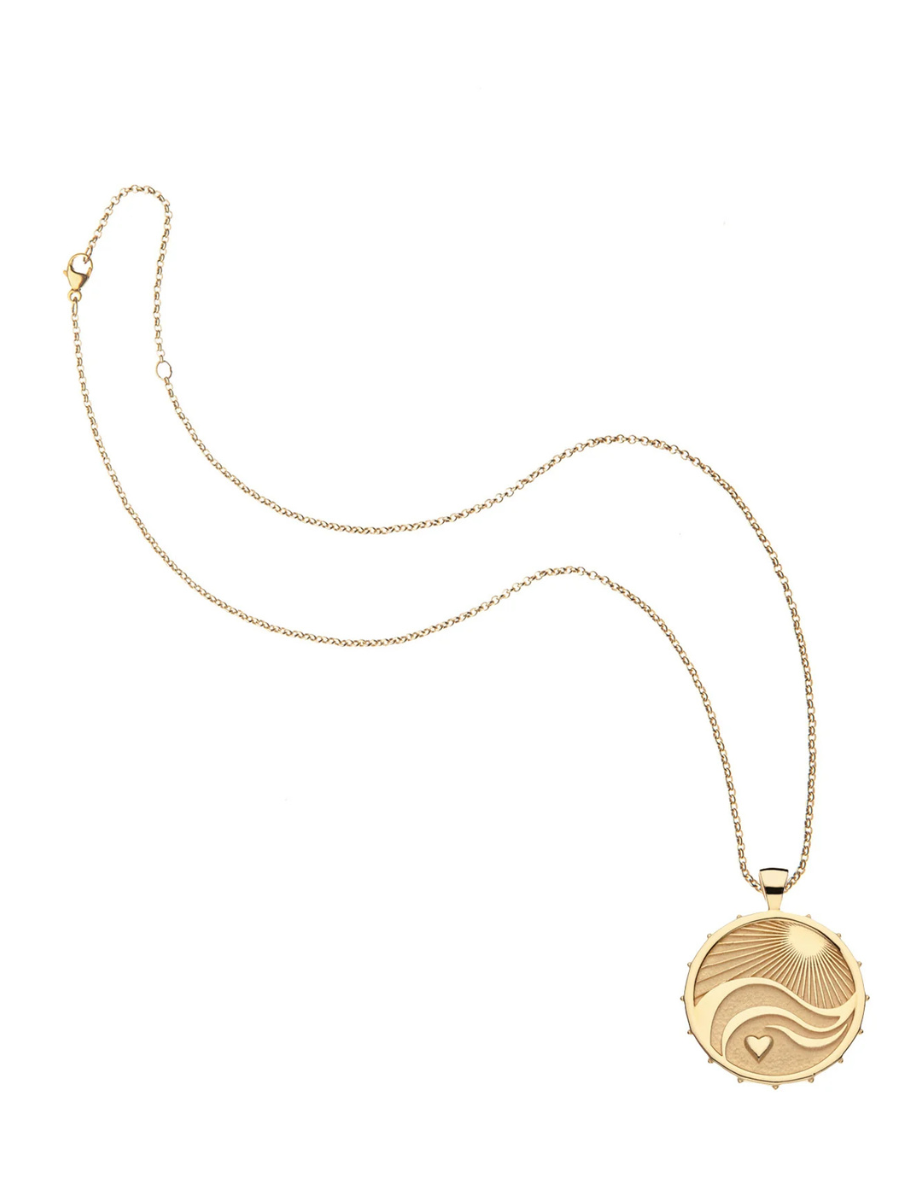 A gold pendant necklace featuring a circular medallion with a sunburst and wave design, displayed on a clean white background.