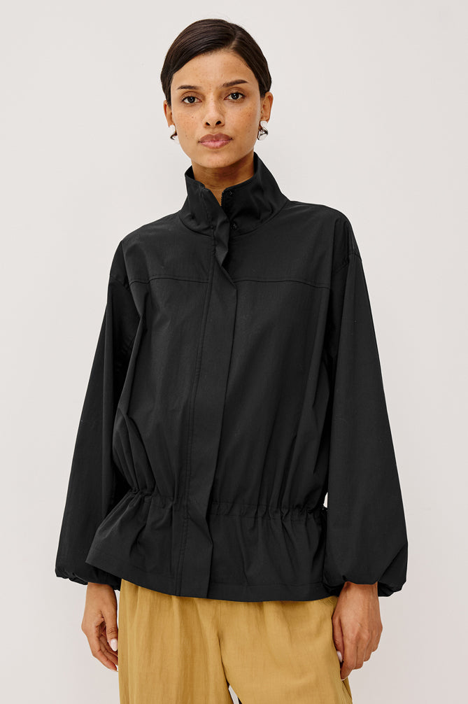 A model wearing a black long-sleeve jacket with a high collar and gathered waist, paired with tan trousers, against a light gray studio background.