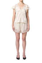 A model wearing a cream-colored two-piece loungewear set consisting of a short-sleeve button-up top with ruffle details and matching lace-trimmed shorts, paired with black heels against a plain white background.