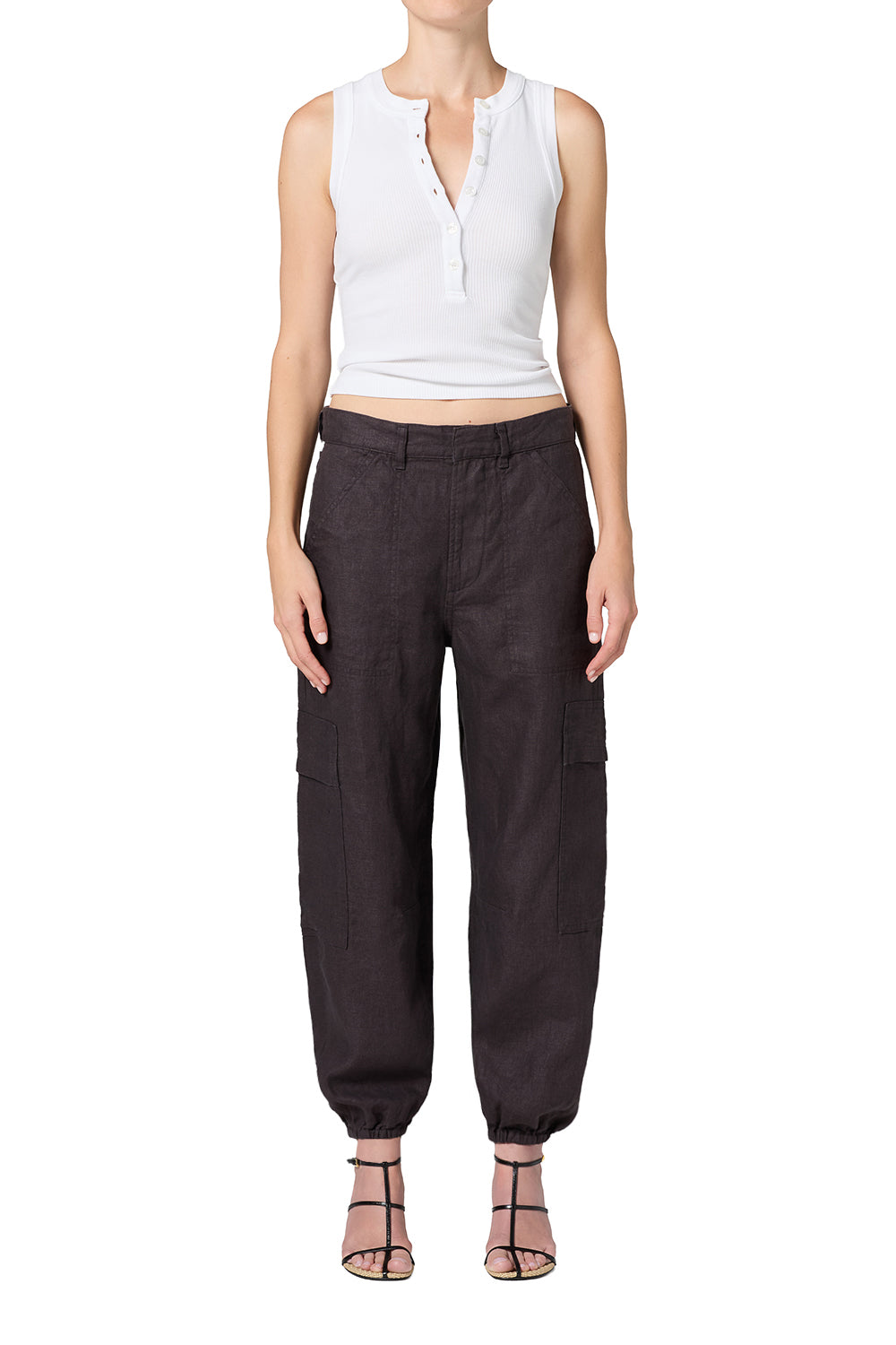 A model wearing dark brown baggy trousers with side cargo pockets, paired with a white sleeveless crop top and strappy heels, shown against a plain white background.