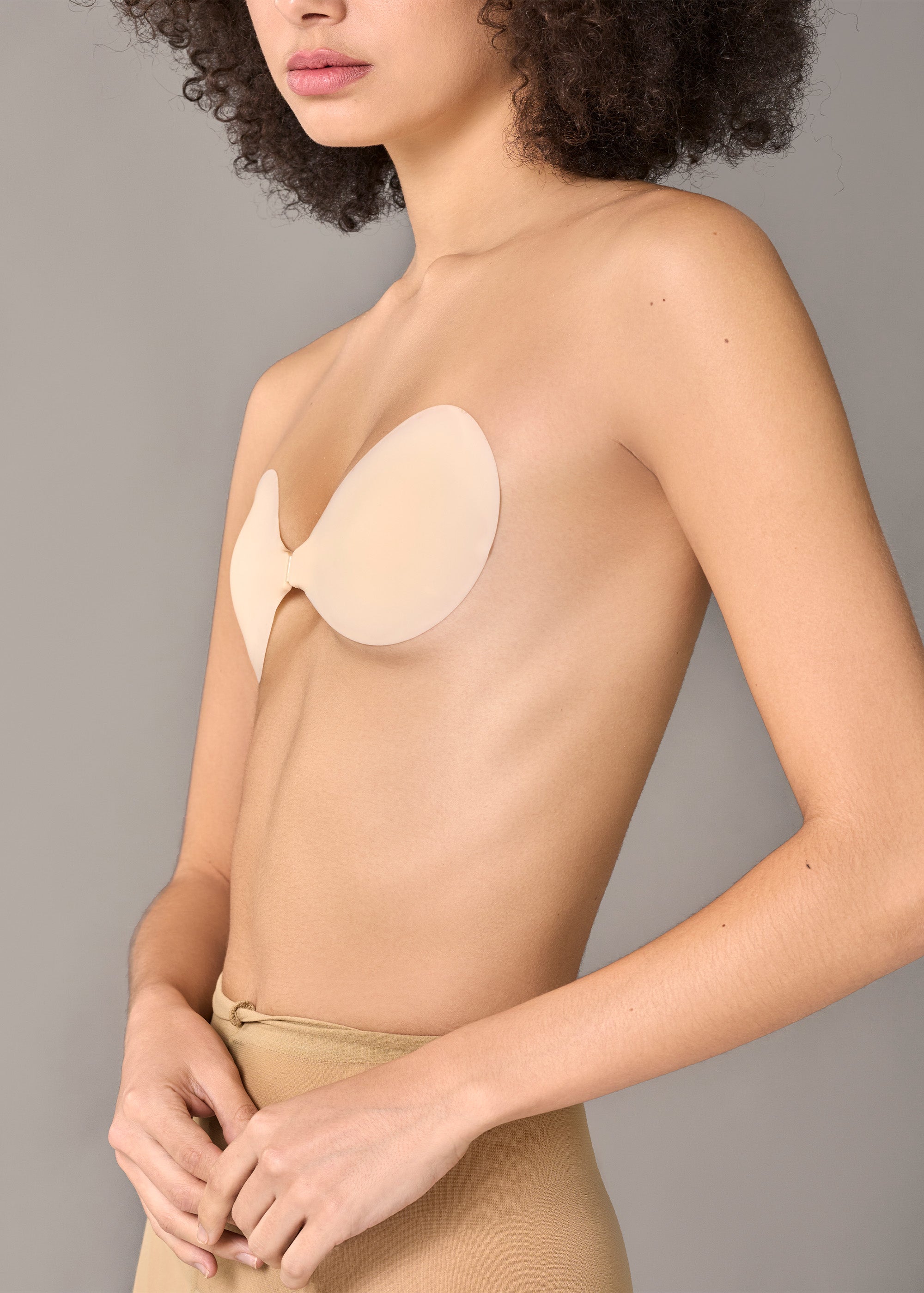 Close-up side view of a woman wearing a light beige bra and matching underwear against a plain gray studio background.