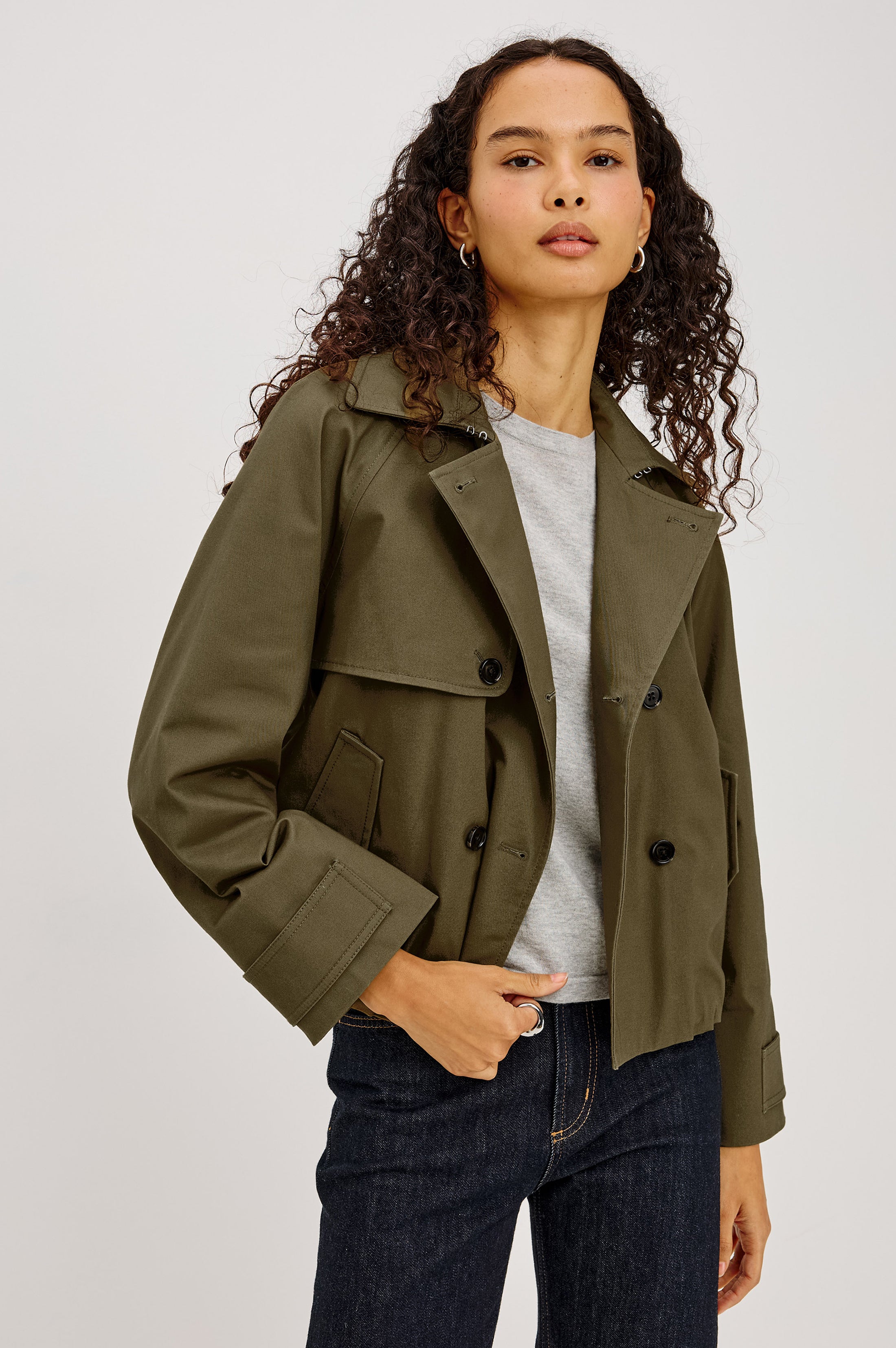 A woman wearing an olive green cropped double-breasted jacket over a light gray t-shirt and dark blue jeans, photographed against a neutral light gray studio background.
