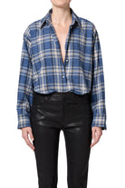 A model wearing a blue, white, and gray plaid button-down shirt tucked into black leather-look pants, shown from the neck down on a white background.