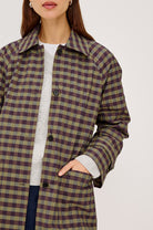A woman wearing a brown and olive green plaid button-up coat over a white knit sweater and dark blue jeans, shown from the chin down against a light gray studio background.
