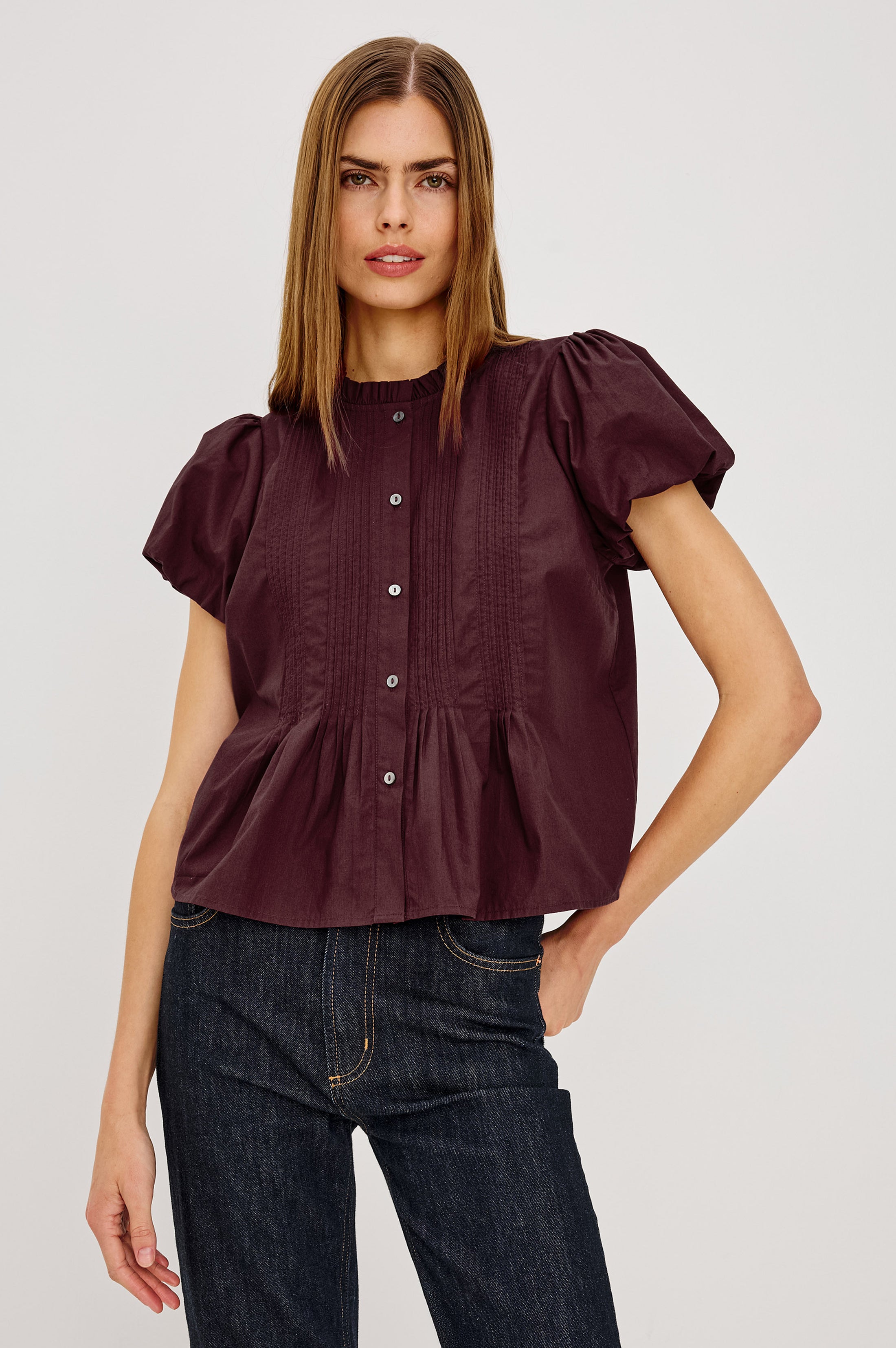 Woman wearing a burgundy blouse with puff sleeves and dark jeans on a light gray background