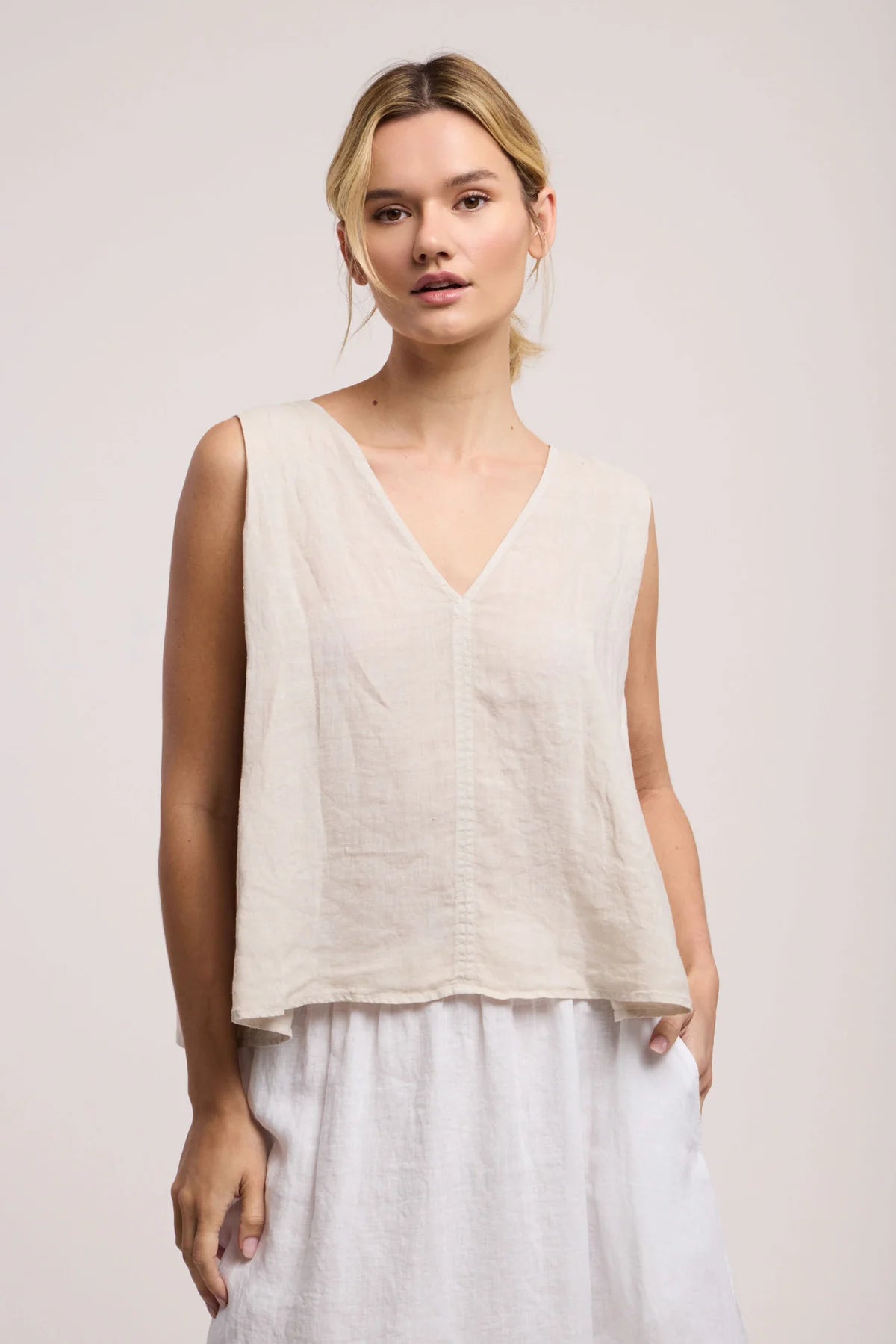 Woman wearing a beige sleeveless top and white pants against a plain background