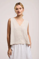 Woman wearing a beige sleeveless top and white pants against a plain background