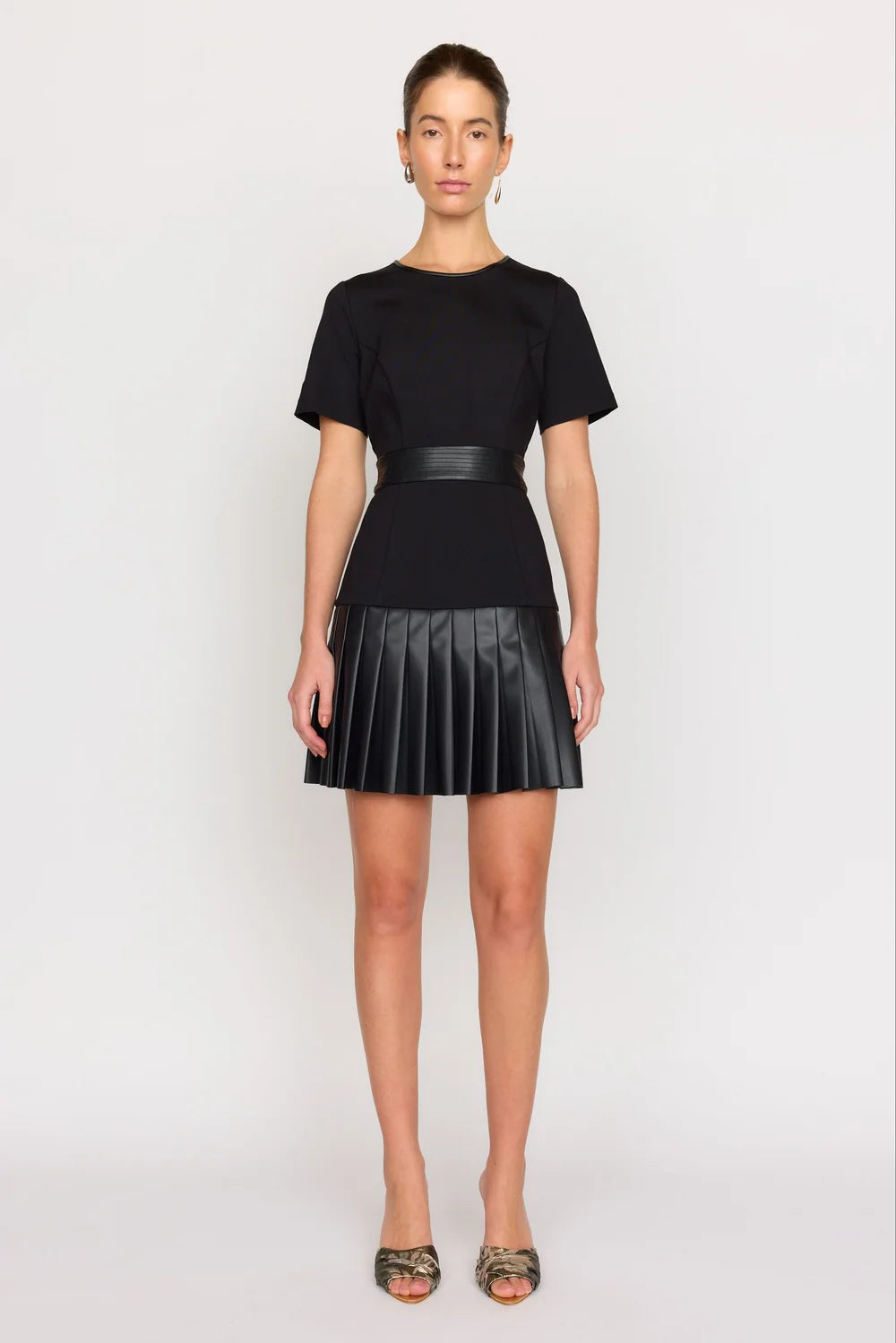 A model stands against a light gray studio background wearing a black short-sleeve top with a matching black pleated mini skirt and a thin black waist belt.